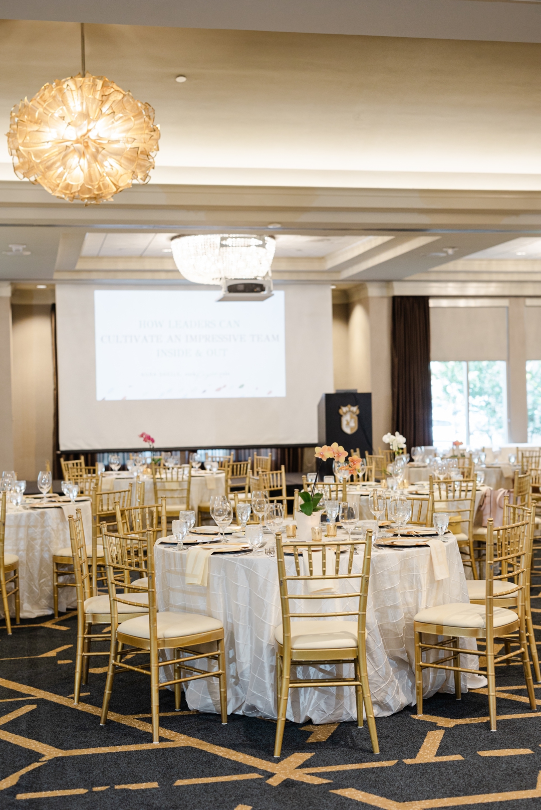 Dallas Event Photographer; A detail photo of a table decorated with a tablecloth, glasses, plates, napkins, and a floral centerpiece. The table is surrounded by gold dining chairs wih cream cushions and the background contains more similar tables with a stage, podium, and projection screen.