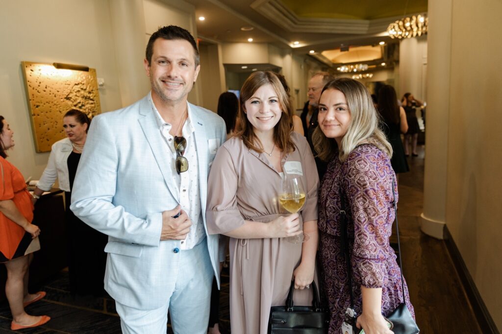 Three members of the Society of Wedding Professionals smiling and posing for the camera. The man on the left is wearing light blue suit and is holding his lapel. The woman in the middle is wearing a tan dress while holding a glass of white wine. And the woman on the right is wearing a purple, floral dress.