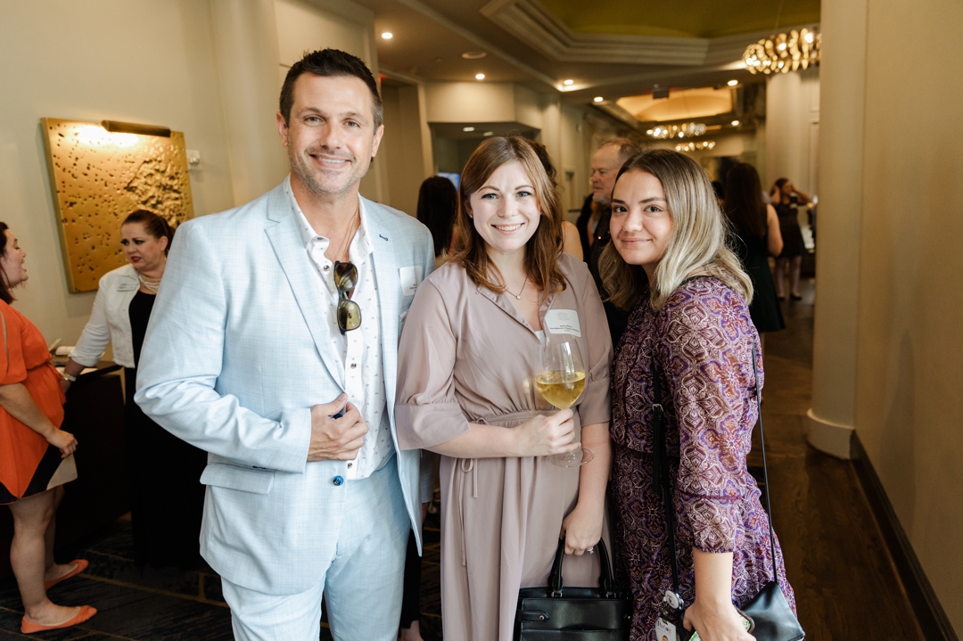 Dallas Event Photographer; Three members of the Society of Wedding Professionals smiling and posing for the camera. The man on the left is wearing light blue suit and is holding his lapel. The woman in the middle is wearing a tan dress while holding a glass of white wine. And the woman on the right is wearing a purple, floral dress.