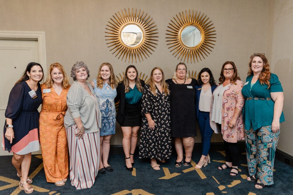 A group of ten woman who are all members of the Society of Wedding Professionals smiling and posing for a group photo. They are all wearing a wide assortment of clothing.