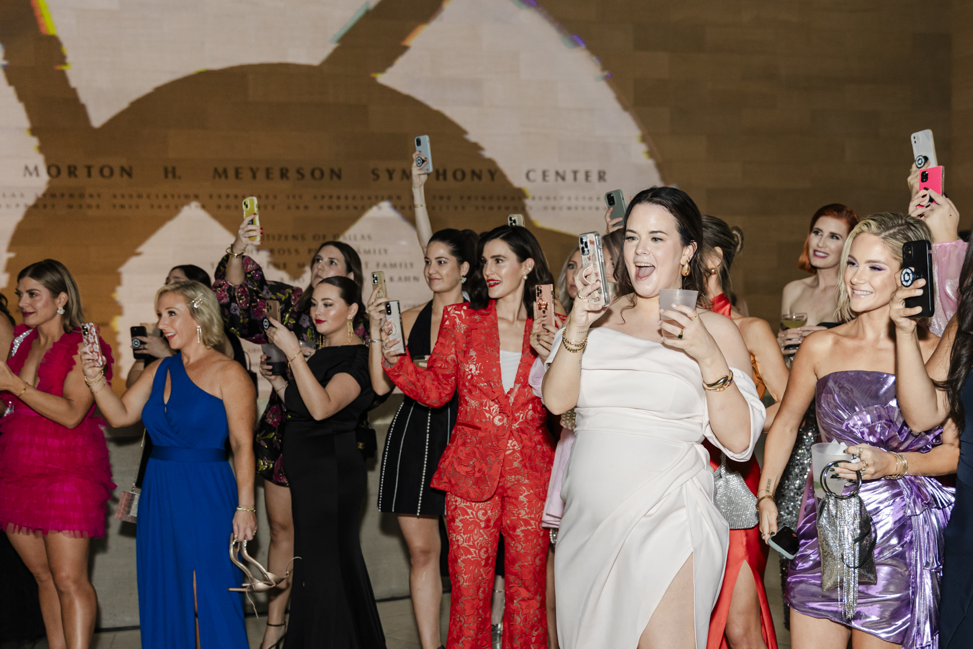 Dallas event photographer capturing a group of women all smiling with their phones out to photograph something out of frame during an event at the Meyerson Symphony Center in Dallas, Texas. All the women are smiling and are wearing formal or cocktail attire.