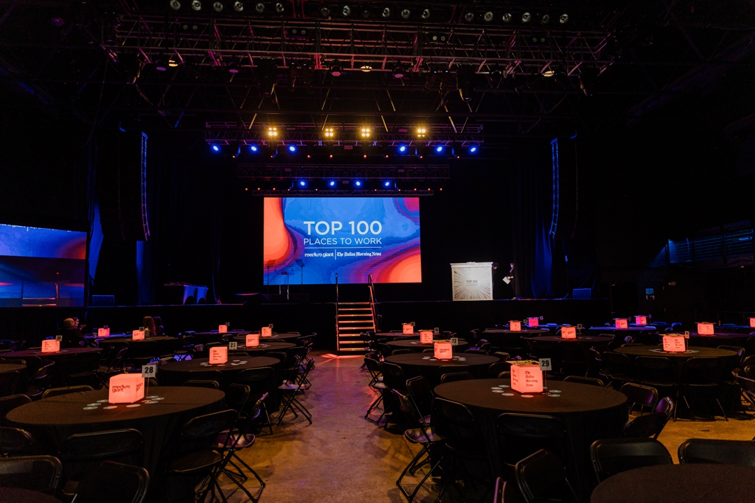 Dallas Event Photographer; A wide shot of the event venue, The Factory in Deep Ellum, with multiple event tables and centerpieces in the foreground and the stage, DJ booth, and projection screen in the background