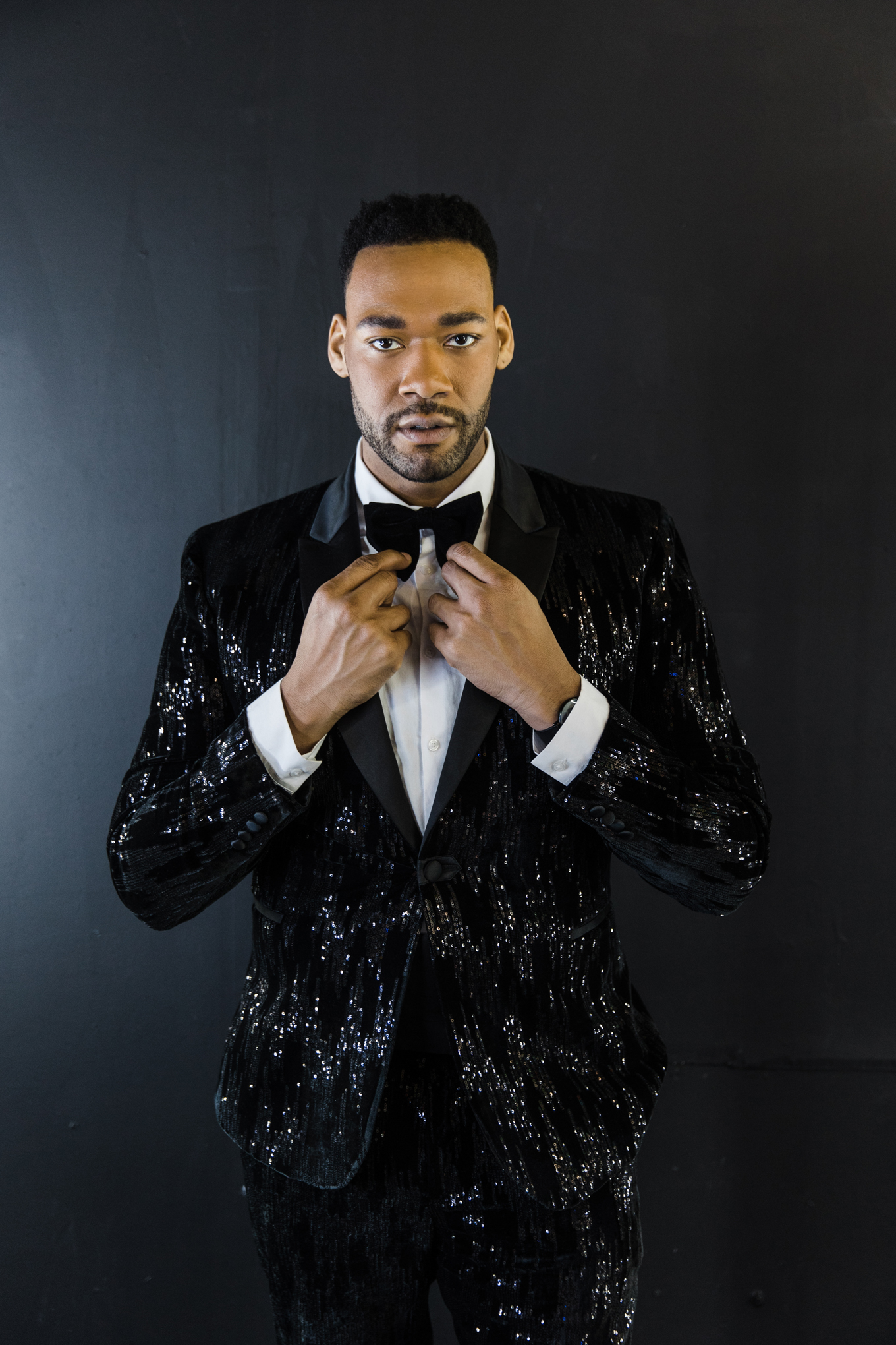 Dallas Fashion Photography; photo of an African American man wearing a sequin tuxedo, adjusting his bow tie, and staring intently at the camera in front of a black background