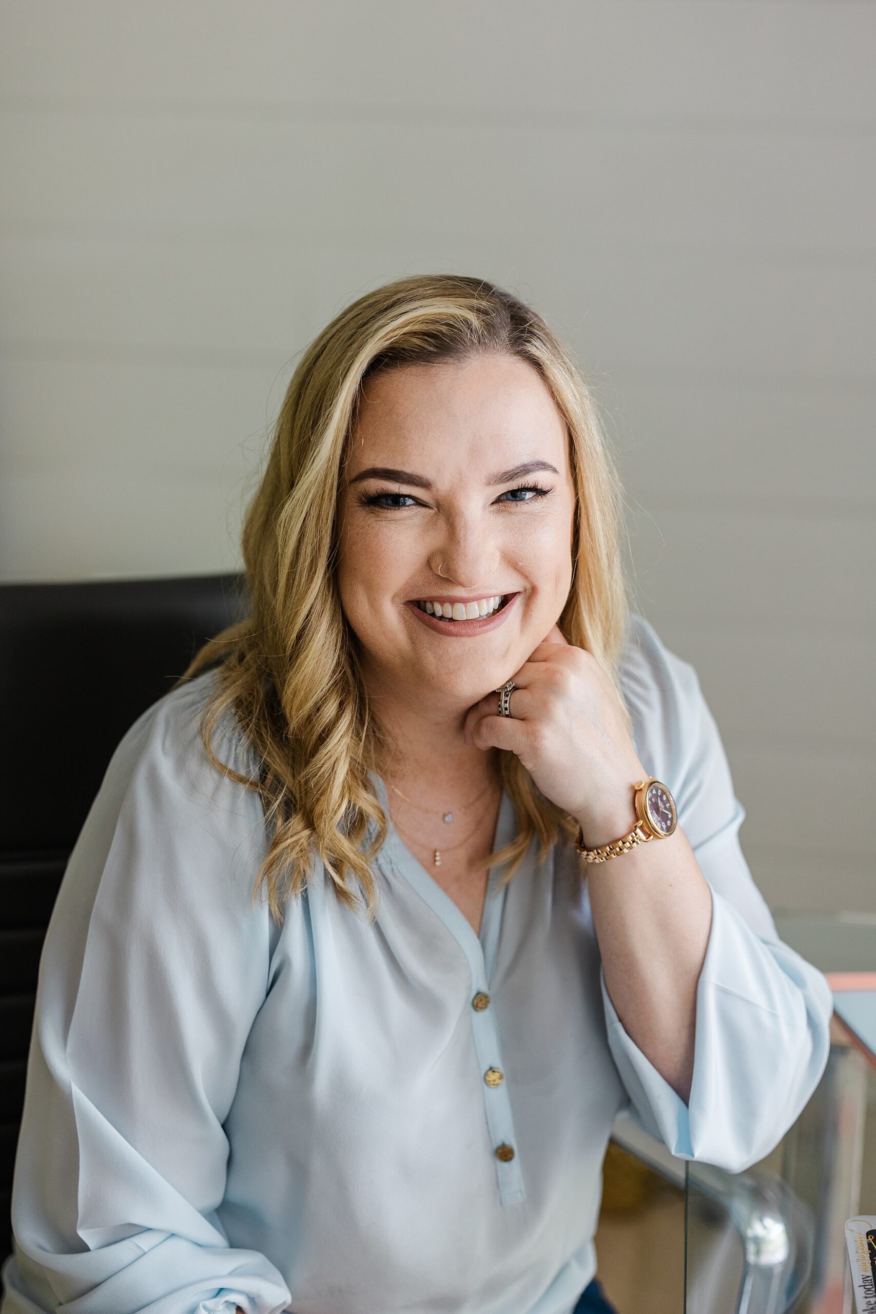 Dallas Fort Worth Author Photographer; Blonde Caucasian woman smiling and looking at the camera while resting her chin on her left hand. She's wearing a light blue top, a gold watch, a silver ring, and a gold necklace. She is sitting on a black chair in front of a white background.