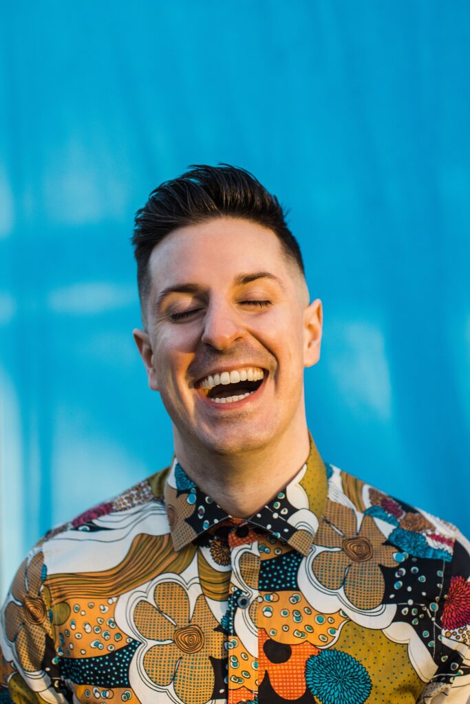 Caucasian male joyfully laughing with his eyes closed in front of a blue background. He has short dark hair and is wearing a highly detailed/colorful floral dress shirt.