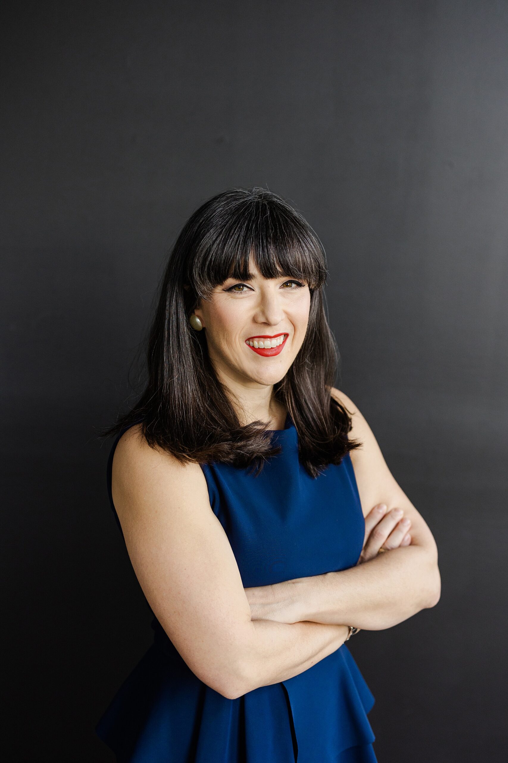 Dallas Fort Worth Author Photographer; Caucasian woman smiling and looking at the camera with her arms folded. She has long, dark hair and is wearing a sleeveless blue top and silver earrings and is in front of a black background.