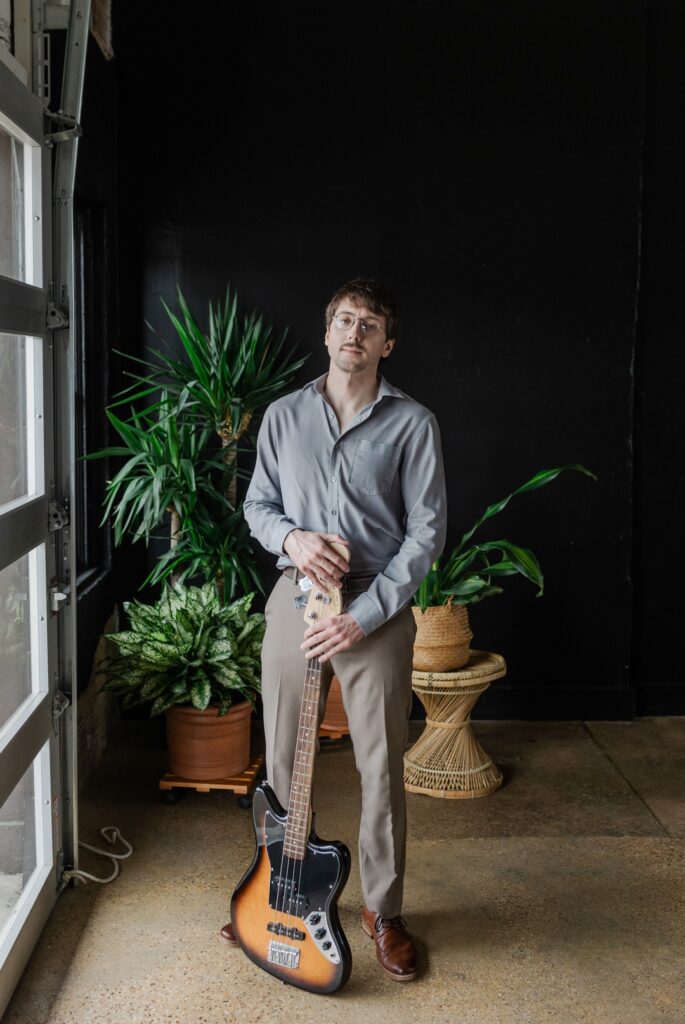 Caucasian man wearing glasses, a grey button down shirt, brown pants, and brown dress shoes, holding a bass guitar and standing next to a window in front of plants and a black backdrop