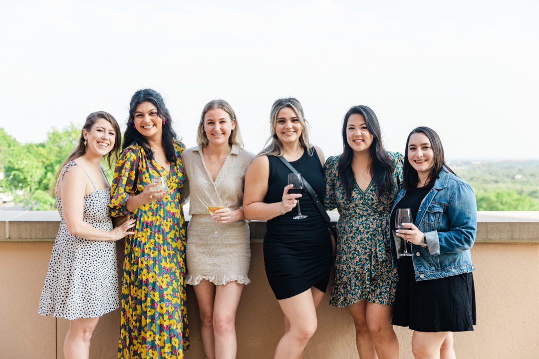 Dallas Corporate Event Photography photo of six women in business casual attire holding drinks, smiling, and posing for the camera at an outdoor event.