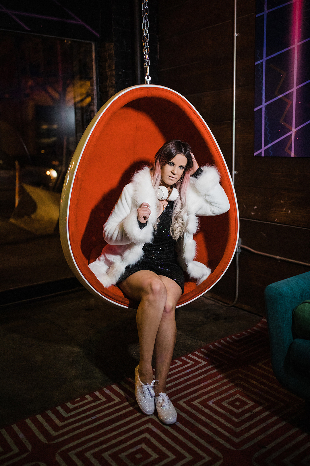 Dallas Fort Worth DJ Photographer; a woman with pink hair highlights wearing sparkly shoes, short black dress, white jacket, and headphones around her neck sitting in a hanging egg-shaped chair in a downtown bar