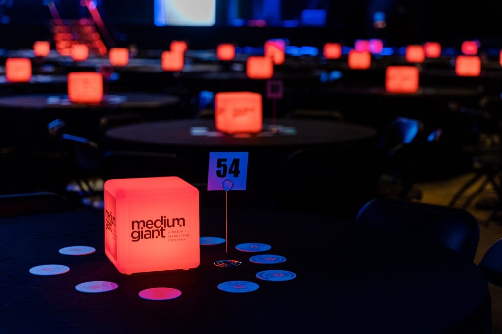 A close up photo of one of the event tables featuring a table number, drink coasters, and a light up cube centerpiece featuring the company, medium giant