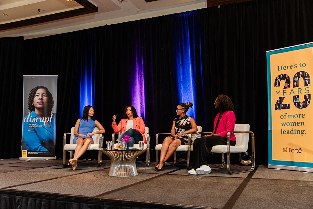 Dallas Fort Worth Event Photographer: four women sitting around a table on stage and having a discussion during the Forte Foundation's MBA Women's Leadership Conference in Dallas, Texas. All four women are wearing professional business attire and are book-ended by tall banners endorsing Forte and their message.