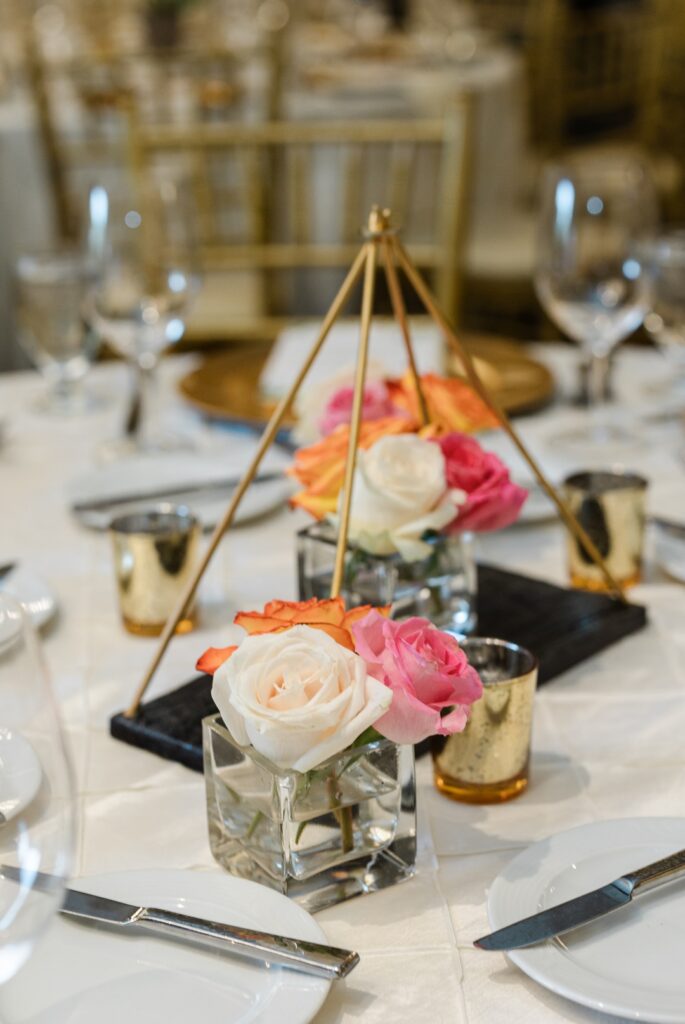 A close up detail shot of a center piece containing white, pink, and orange flowers in a glass cube container. A second glass cube with similar flowers is contained in a golden pyramid.