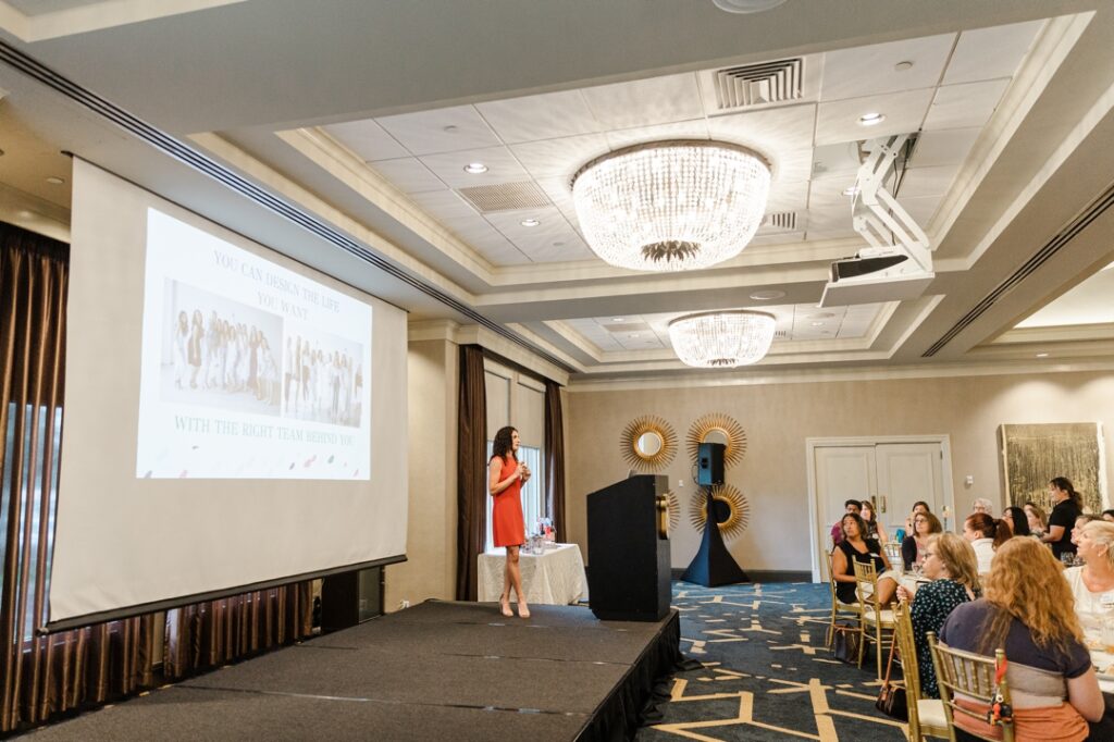 A member of the Society of Wedding Professionals in an orange dress on stage giving a presentation with a projection screen and podium to a crowd sitted at tables.