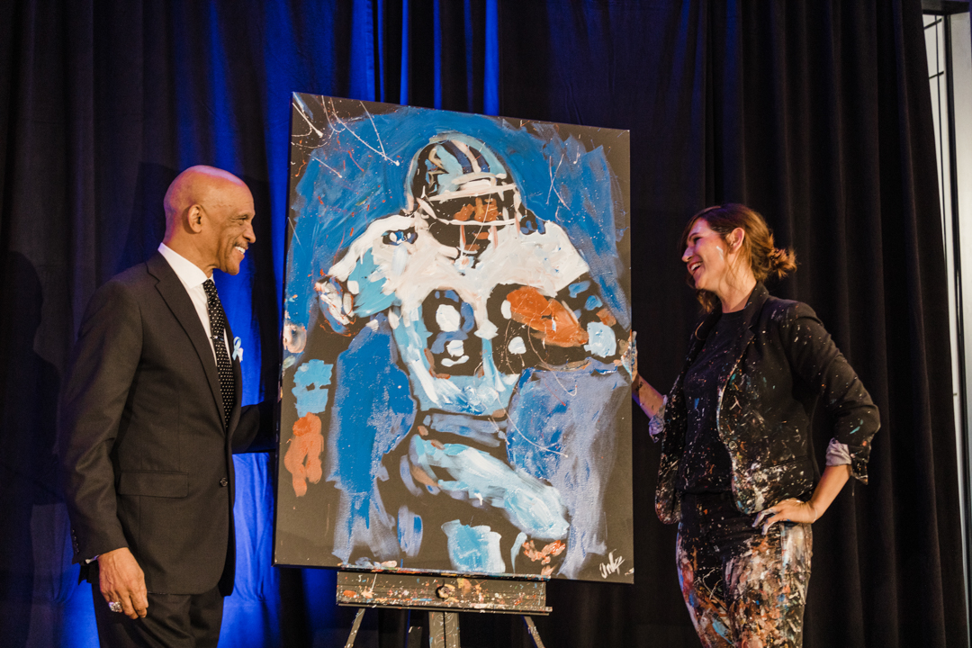 Dallas Fort Worth Fundraiser Event; photo of a man and woman auctioning off a football painting on a stage at a fundraising event.