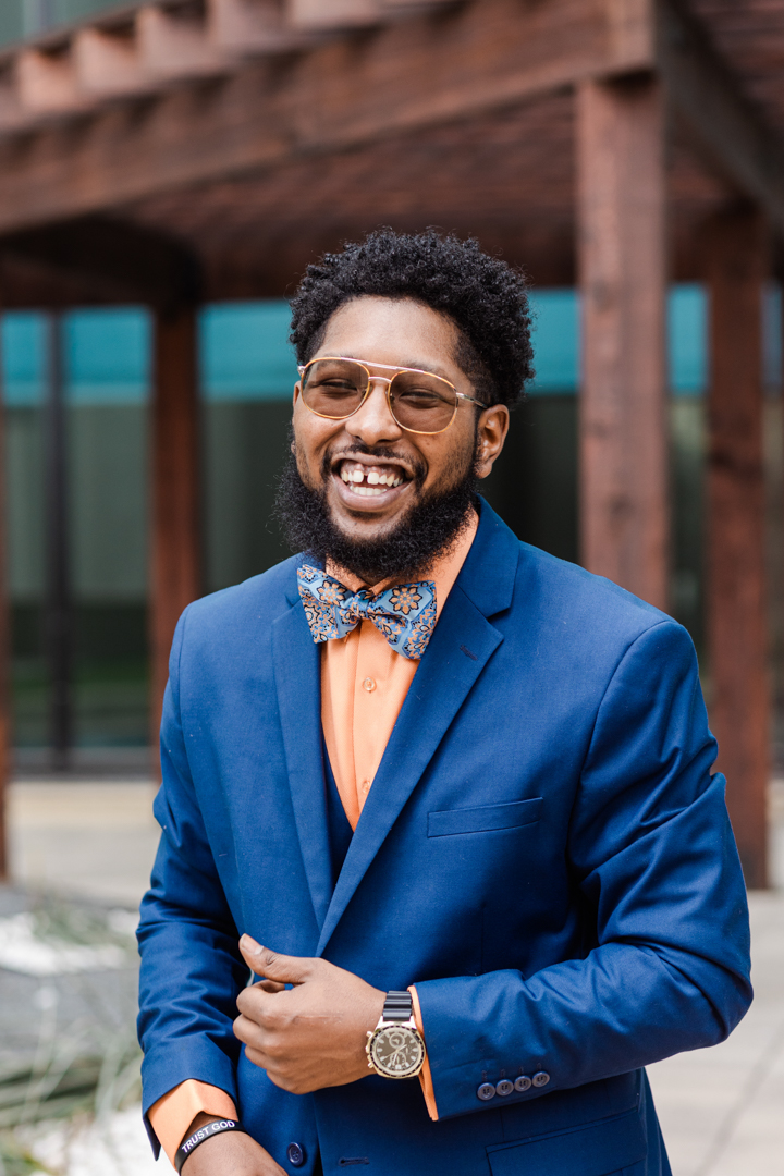Dallas Fort Worth Headshot Photographer; photo of a man in a blue and orange suit with a bowtie and glasses laughing and holding his lapel in front of an outdoor setting