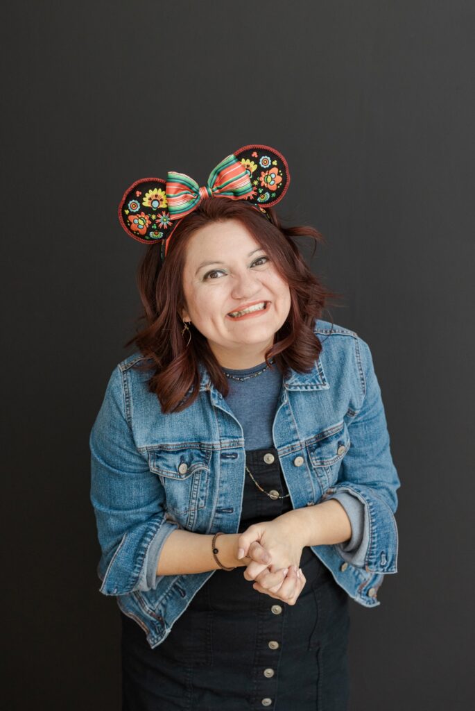 Latina woman wearing denim and a colorful mouse ear bow clasping her hands and smiling in front of a dark background