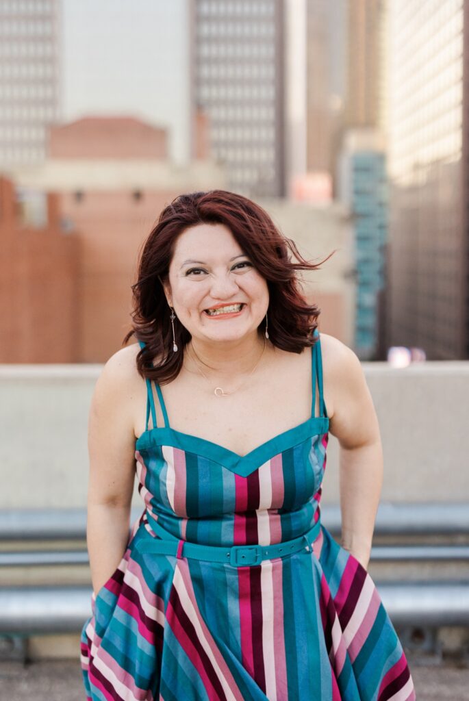 Latina woman wearing a colorfully striped dress with a blue belt smiling and posing on top of a parking garage in front of a city skyline