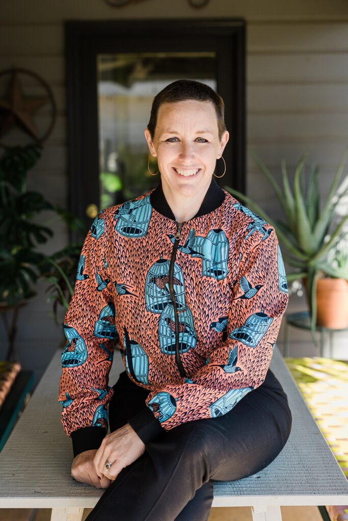 Detail shot of a Caucasian woman's top. The top is orange with blue birds and blue bird cages all over the top alongside many small black decorations. The woman's face is cutoff but her smile is partially seen. In the background are many different plants.
