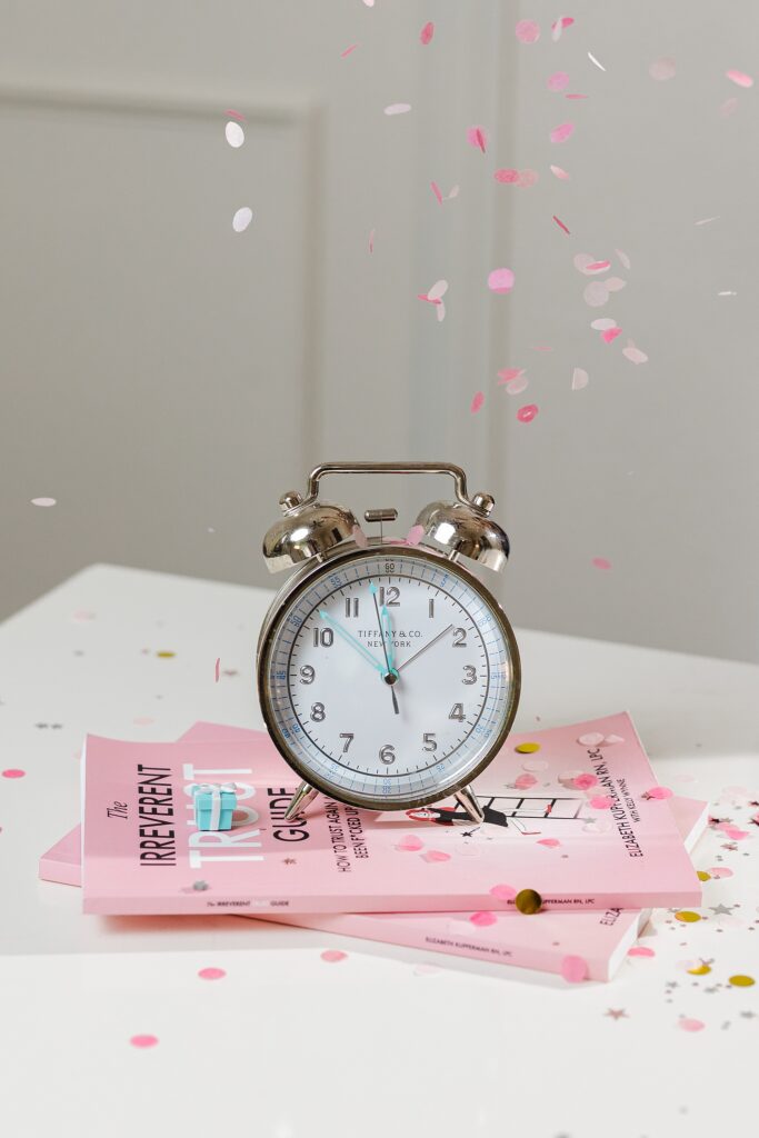 Detail shot of a Tiffany & Co. traditional alarm clock. The clock has blue hands and has confetti around it on multiple sides. The clock is resting on a pink book on top of a white desk.