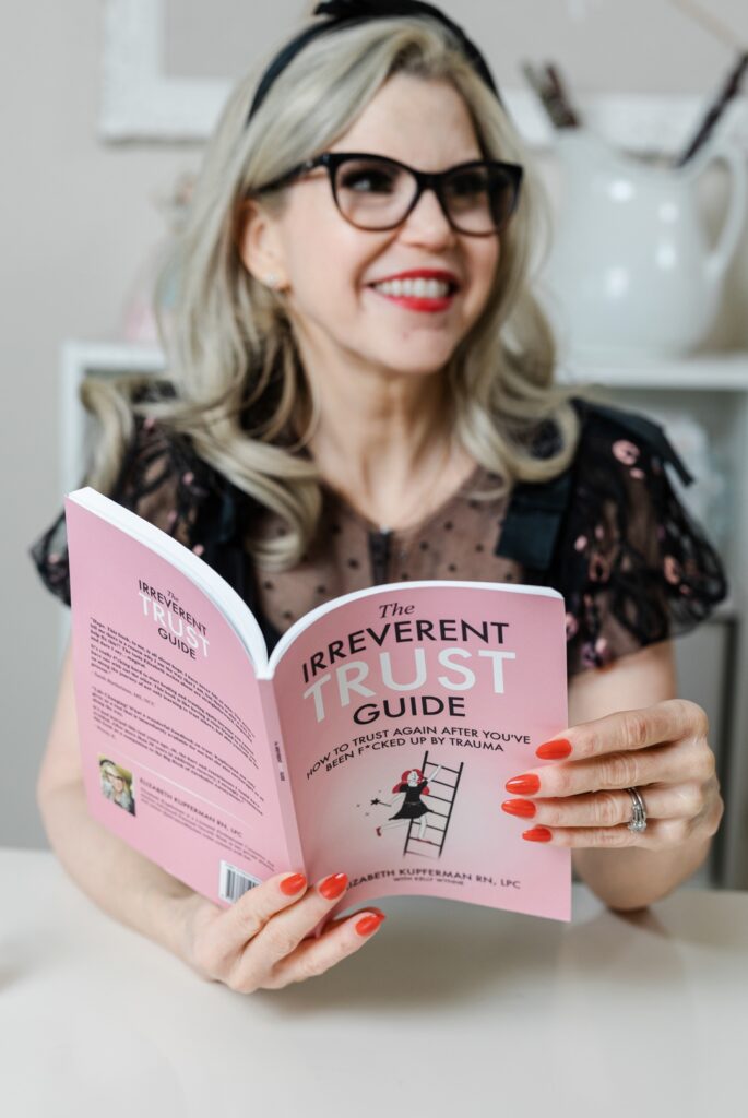 Smiling, caucasian woman wearing a black blouse, black glasses, and a black headband looking off into the distance while holding an open, pink book titled "The Irreverent Trust Guide" by Elizabeth Kupferman