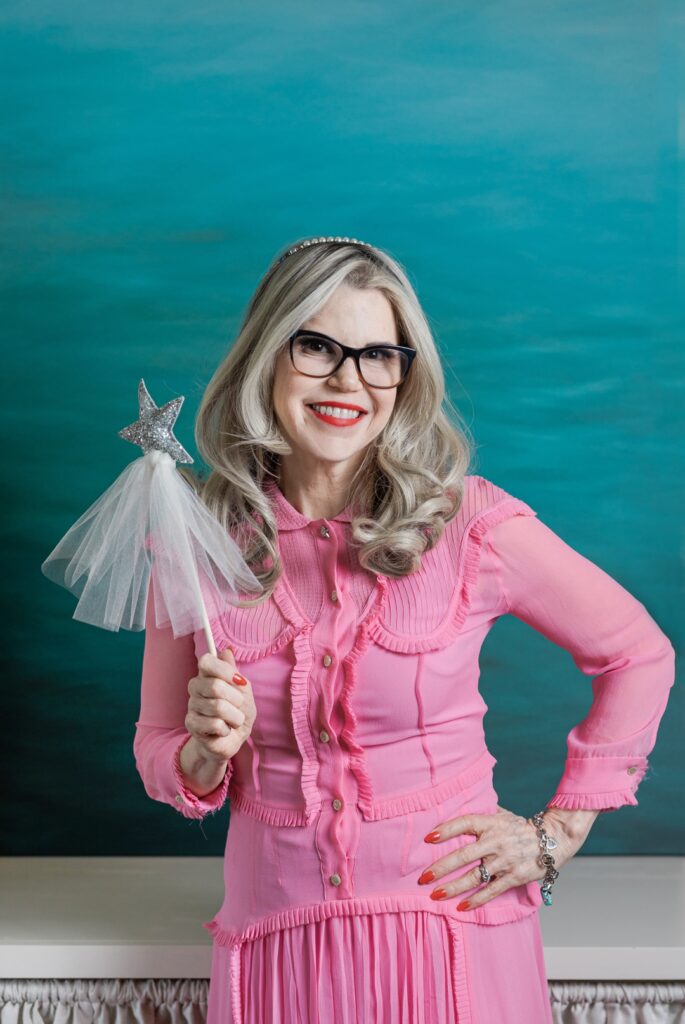 Smiling, caucasian woman wearing a pink dress, black glasses, and a silver headband while standing with one arm akimbo and one arm holding a magic wand while standing in front of a teal backdrop
