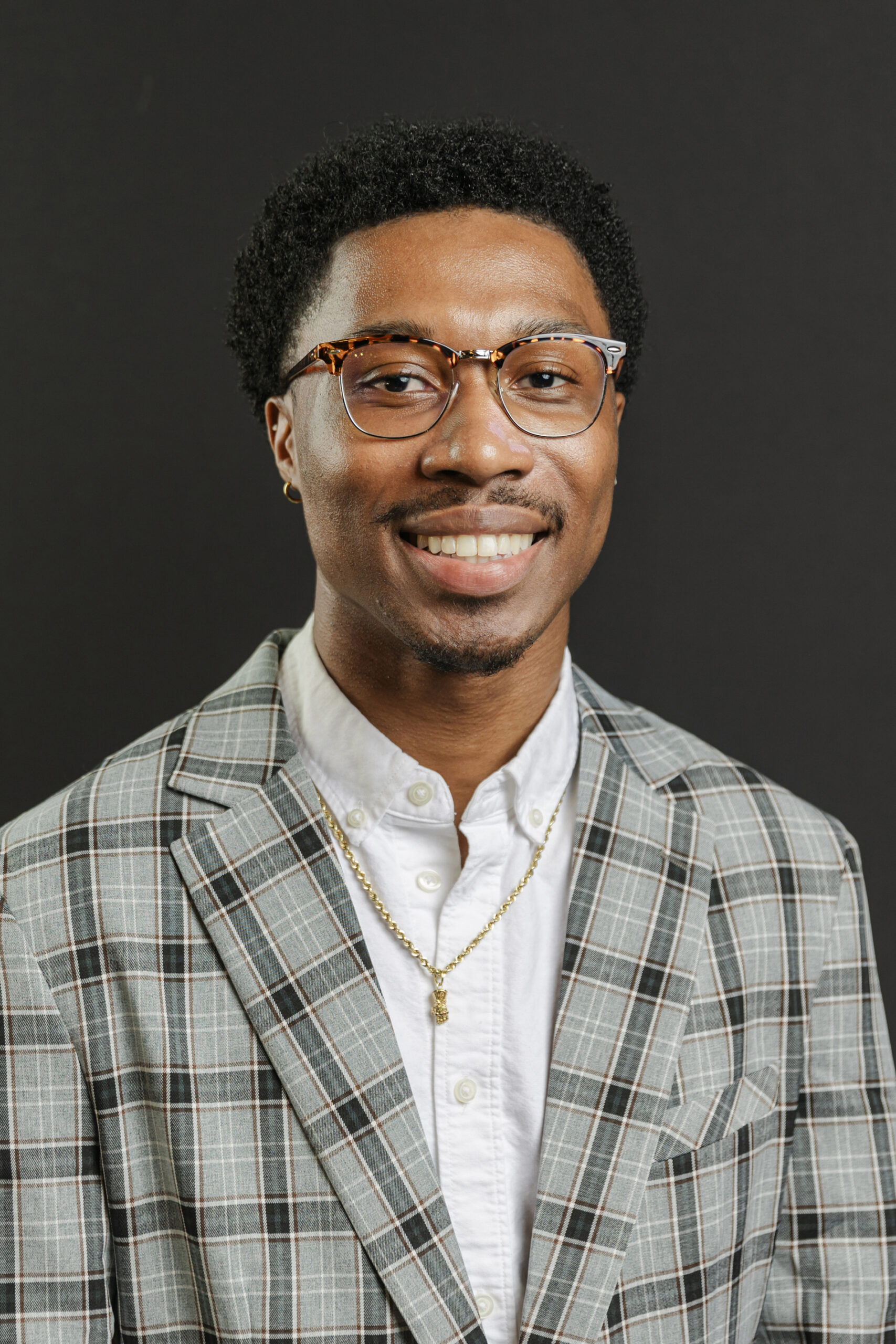 Dallas head shot photographer: head shot of a man in front of black background. He's wearing a white dress shirt, plaid jacket, glasses, and a gold necklace.