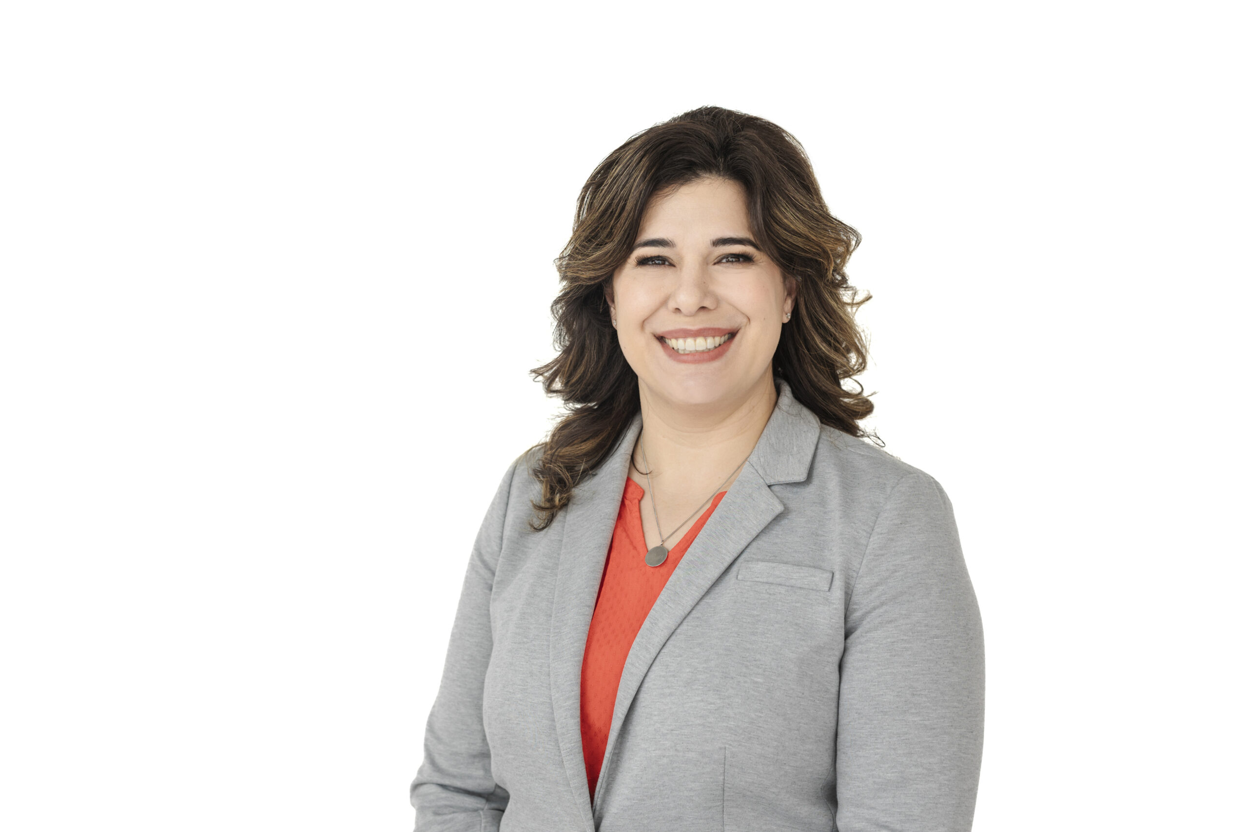Head shot of a woman in front of a white background. The woman is smiling at the camera while wearing an orange top and a gray blazer with a necklace.