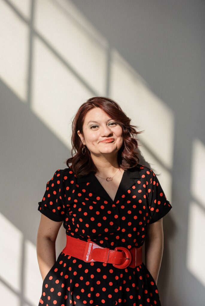 Latina woman wearing a black and red polka dotted dress with a red belt and posing in front of a white background and using the window light to frame her