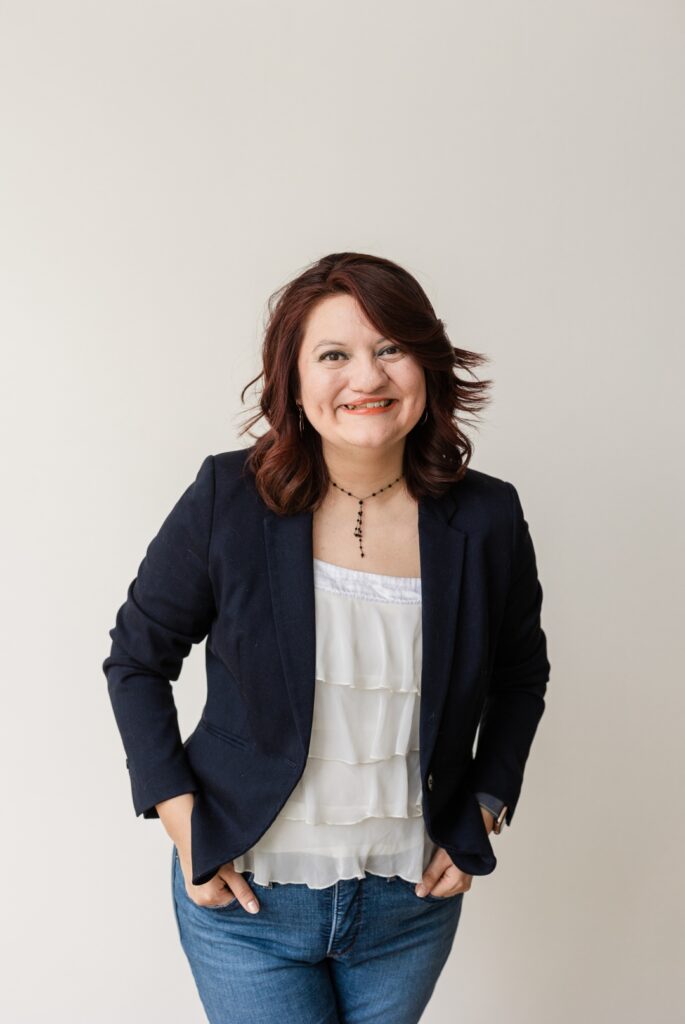 Latina woman wearing jeans, a white blouse, and a dark blazer posing in front of a white background