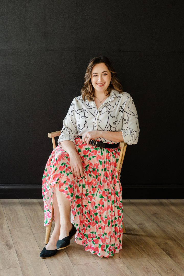 Dallas Headshot Photographer; photo of a woman in a colorful skirt and intricate blouse sitting cross legged in a wooden chair and smiling in front of a black background