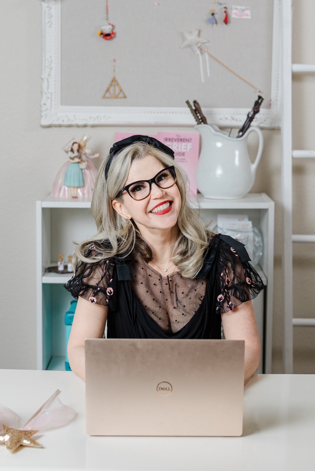 Dallas Therapist Headshot Photographer; Smiling, caucasian woman wearing a black blouse, black glasses, and a black headband sitting behind a laptop and a magic wand