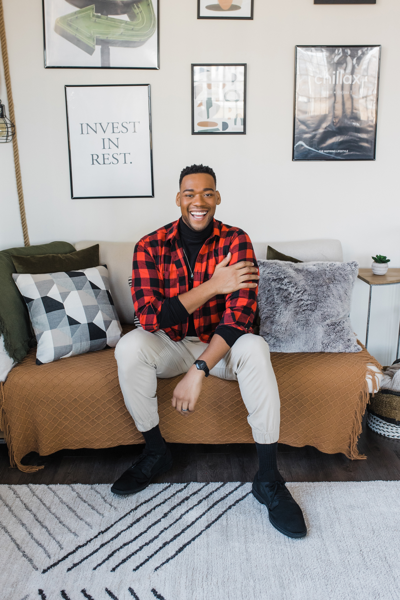 Dallas Lifestyle Brand Photographer; photo of an African American man wearing a black shirt, red flannel jacket, tan pants, and black shoes smiling and sitting on a couch in front of a wall covered in framed art and photos