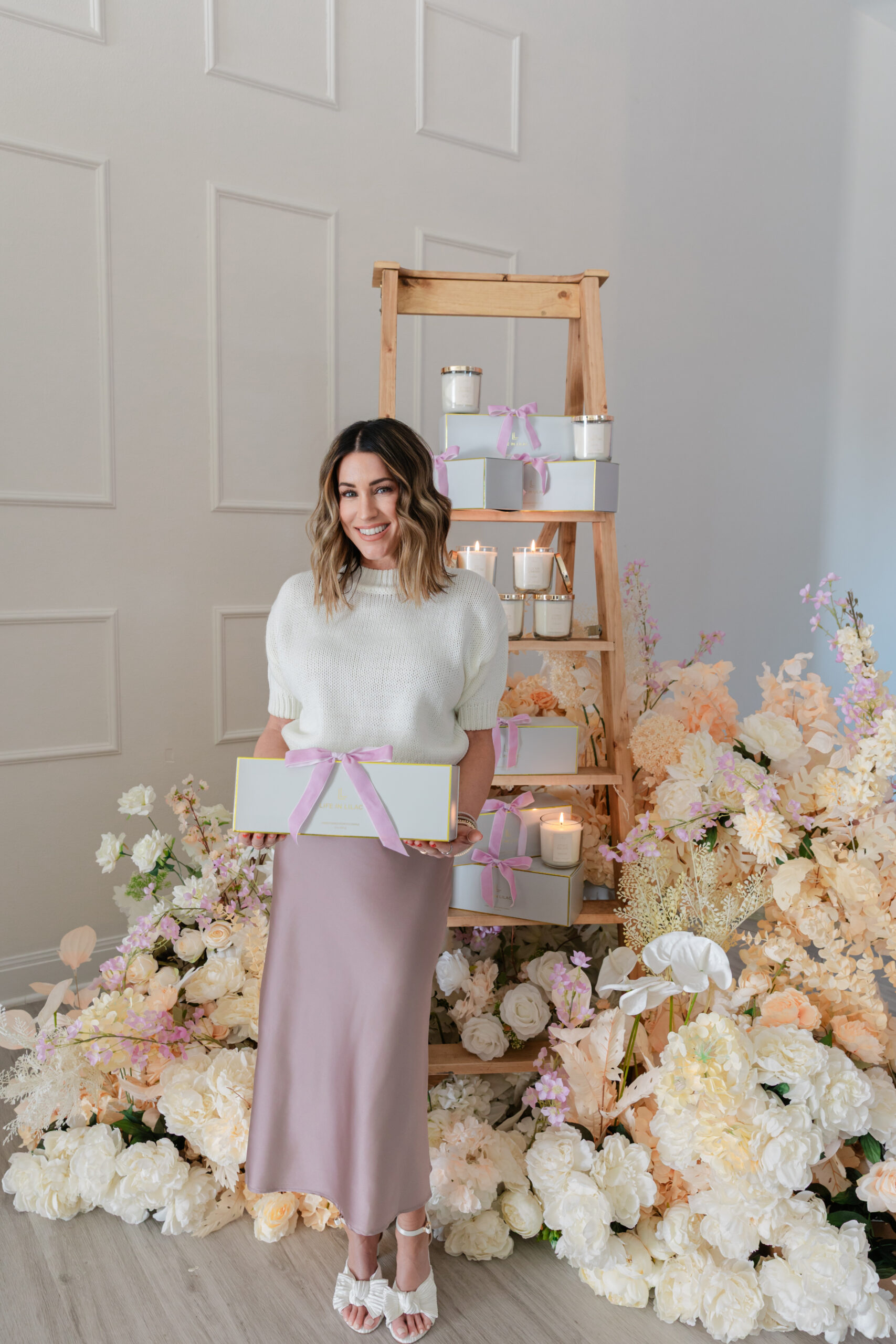 the founder of Life In Lilac holding a box and standing next to her candles, a large floral arrangement 