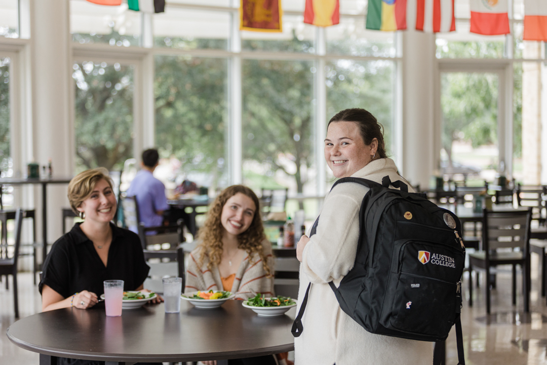 Dallas Lifestyle Photography; photo of three women smiling and preparing to eat lunch in a college setting; one of them is wearing a backpack that shows the logo for Austin College