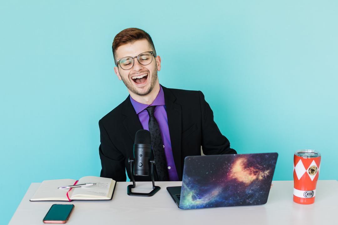 Dallas Podcast Photographer; man in a black suit, purple dress shirt, and black tie laughing while sitting at a desk covered in podcast accessories: microphone, laptop, notebook, thermos, etc. all in front of a light blue background