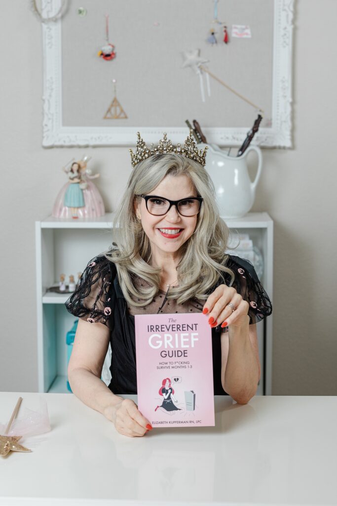 Caucasian woman with blonde hair holding a copy of her book. She's wearing black glasses, silver earrings/rings, and a decorative black top with pink accents. She is in front of a white bookshelf that has dolls, a pitcher, and other objects.