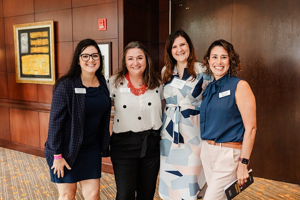 Four women smiling and posing together during the Forte Foundation's MBA Women's Leadership Conference in Dallas, Texas. All four women are dressed in varying types of professional business attire and have on name tags with the Forte logo.