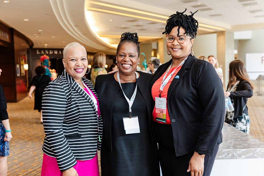 Three women smiling and posing together during the Forte Foundation's MBA Women's Leadership Conference in Dallas, Texas. All three women are dressed in varying types of professional business attire and have on Forte branded lanyards with name tags.