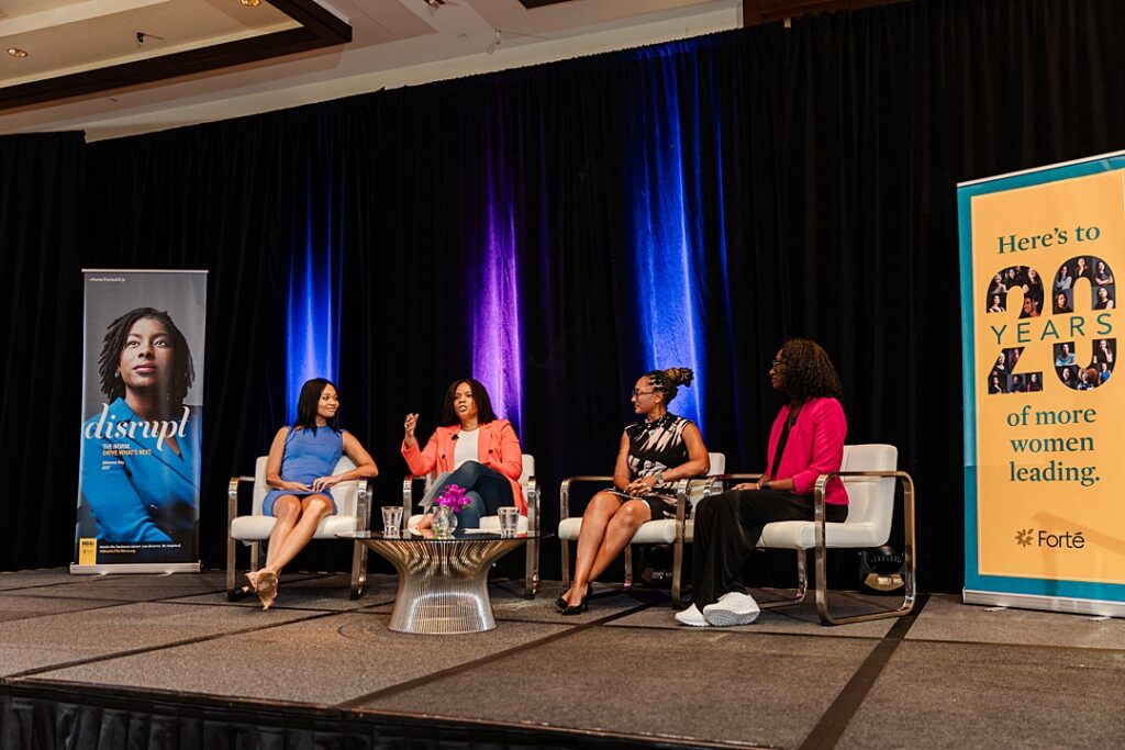Four women sitting around a table on stage and having a discussion during the Forte Foundation's MBA Women's Leadership Conference in Dallas, Texas. All four women are wearing professional business attire and are book-ended by tall banners endorsing Forte and their message.