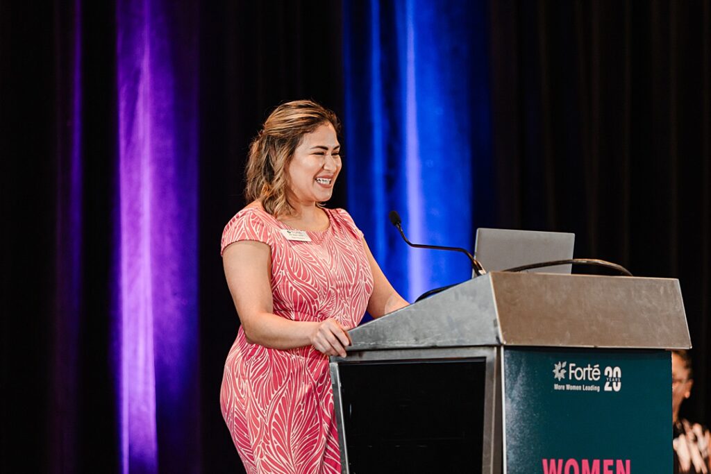 A woman smiling and speaking on stage from a podium during the Forte Foundation's MBA Women's Leadership Conference in Dallas, Texas. She's wearing a intricate, floral, red and white dress and a Forte name tag.