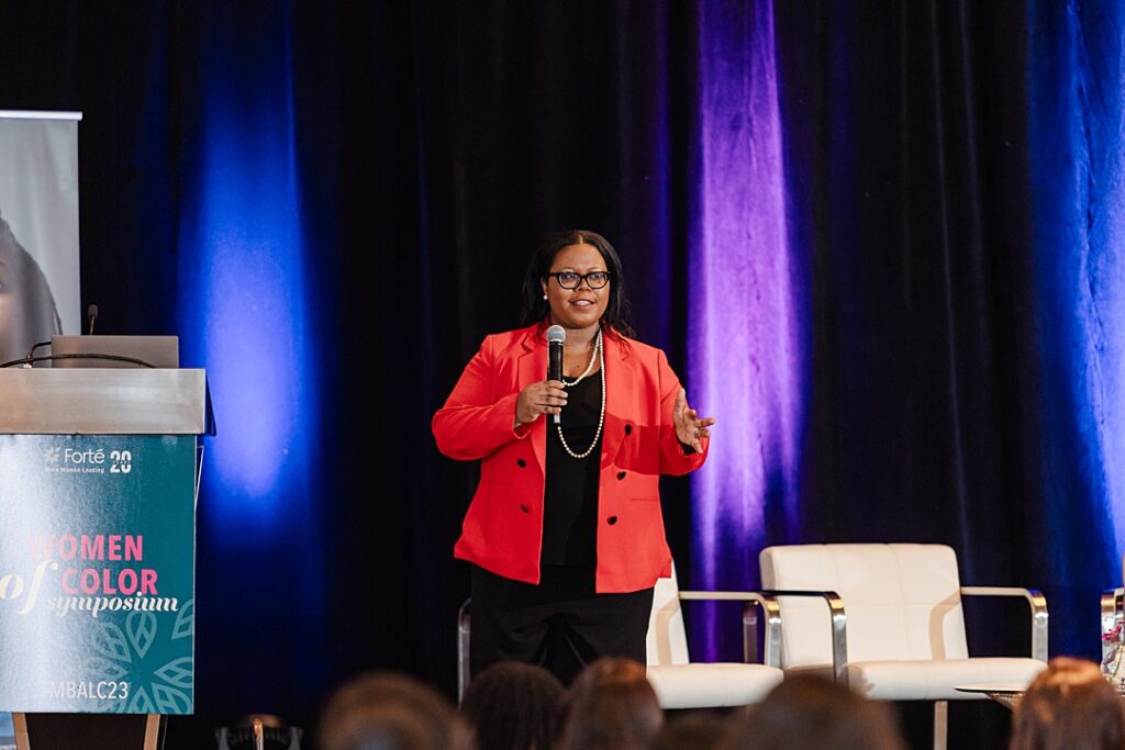 A woman addressing the audience from the stage during the Forte Foundation's MBA Women's Leadership Conference in Dallas, Texas. She's wearing a black top, black pants, and a red jacket with glasses. A podium and chairs can be seen on either side of her.