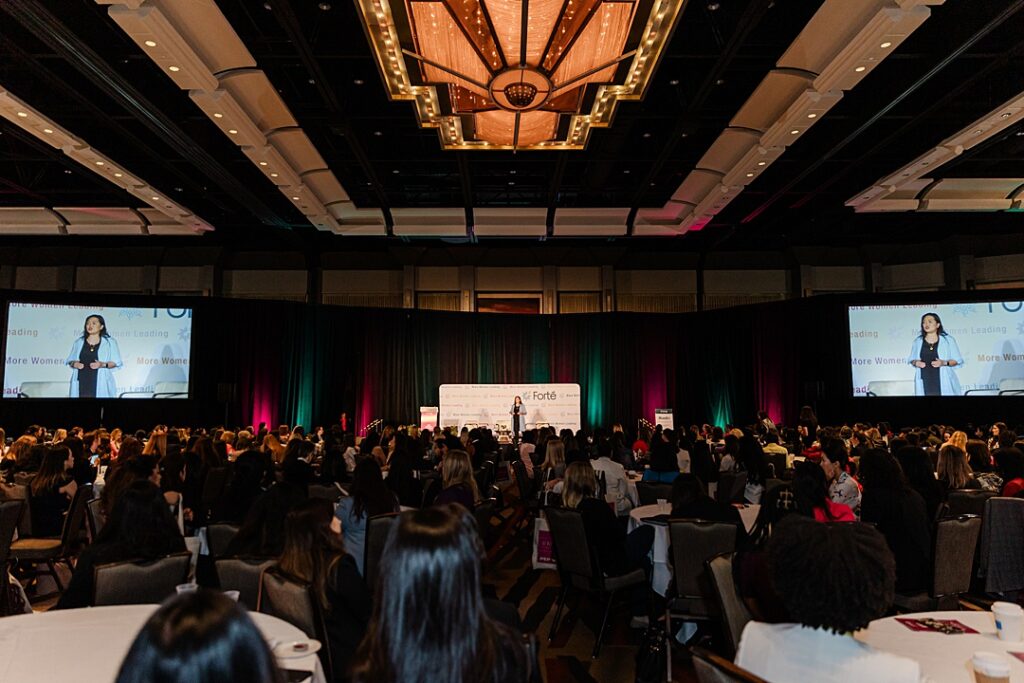A wide shot of a ballroom filled with women and a speaker on stage during the Forte Foundation's MBA Women's Leadership Conference in Dallas, Texas. All the women are sitting at round ballroom tables while the speaker is also being projected onto two large screens on either side of the stage.