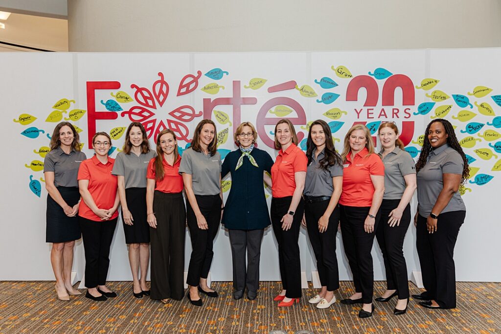 Eleven women posing in front of a inspiration wall during the Forte Foundation's MBA Women's Leadership Conference in Dallas, Texas. Ten of the women are wearing matching polos and black pants while the woman in the middle is wearing professional business attire. The wall behind them reads "Forte 20 Years" and is covered in leaves with motivations written by the conference attendees.