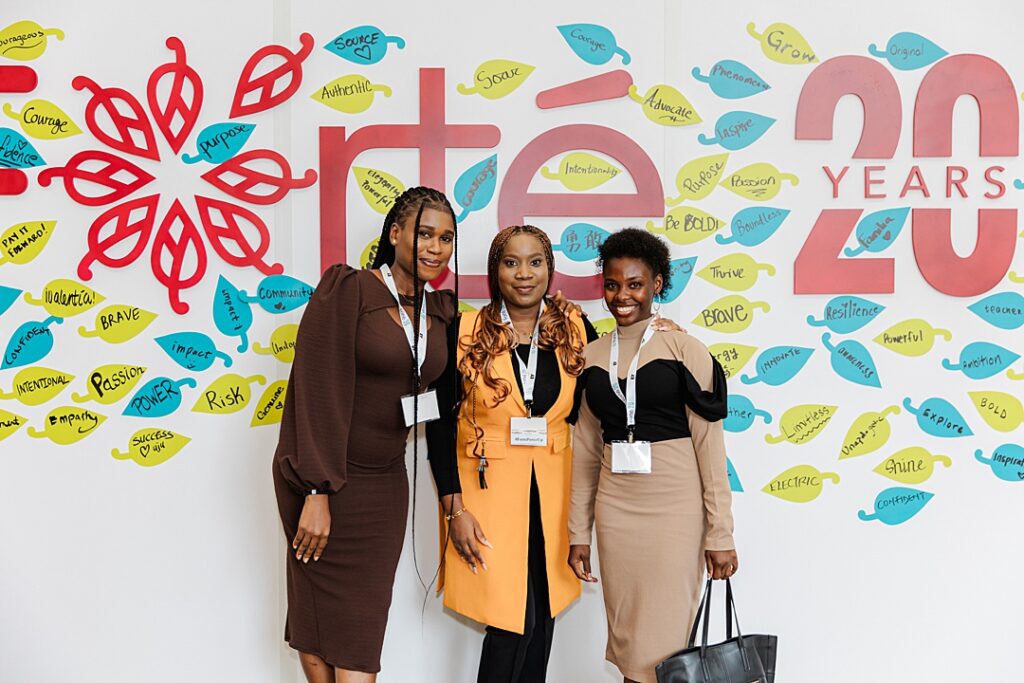 Three women posing in front of a inspiration wall during the Forte Foundation's MBA Women's Leadership Conference in Dallas, Texas. All three women are wearing professional business attire and Forte branded lanyards. The wall behind them reads "Forte 20 Years" and is covered in leaves with motivations written by the conference attendees.