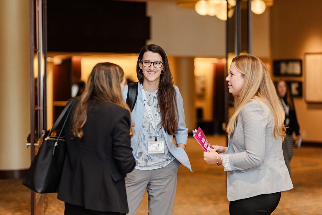 Three women smiling and interacting during the Forte Foundation's MBA Women's Leadership Conference in Dallas, Texas. All three women are wearing professional business attire and Forte branded lanyards.