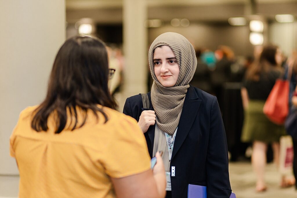 Two women talking during the Forte Foundation's MBA Women's Leadership Conference in Dallas, Texas. Both women are wearing professional business attire and Forte branded lanyards. The woman facing the camera is also wearing a hijab.