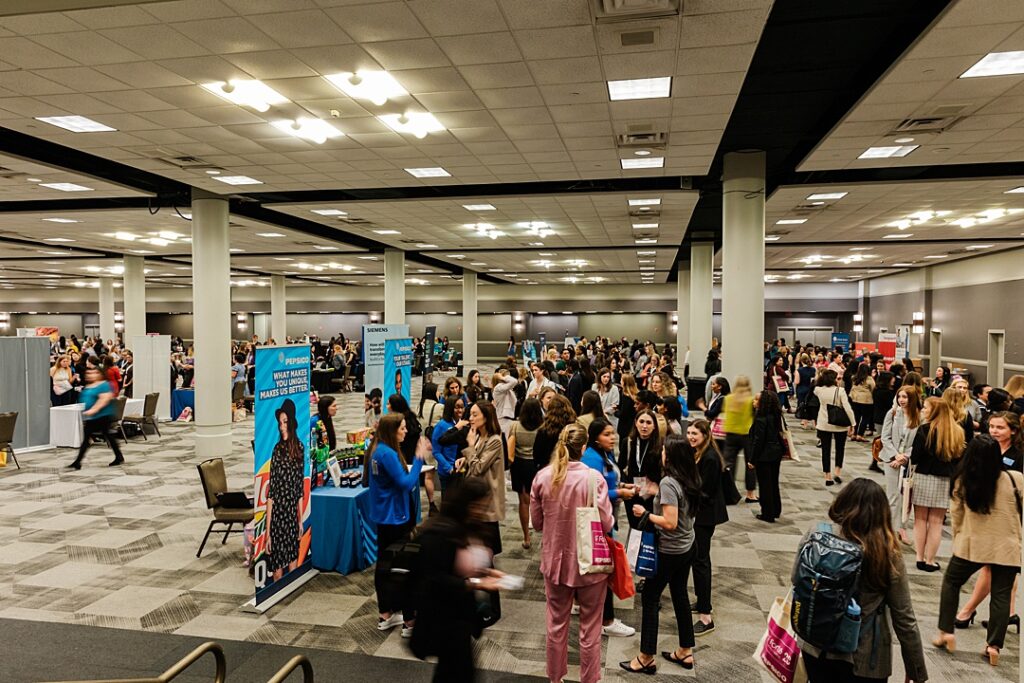 Wide shot of the large trade show that occurred on the last day of the Forte Foundation's MBA Women's Leadership Conference in Dallas, Texas. Rows of tables run by different businesses line the large hall while hundreds of professional women move from one business to the next.