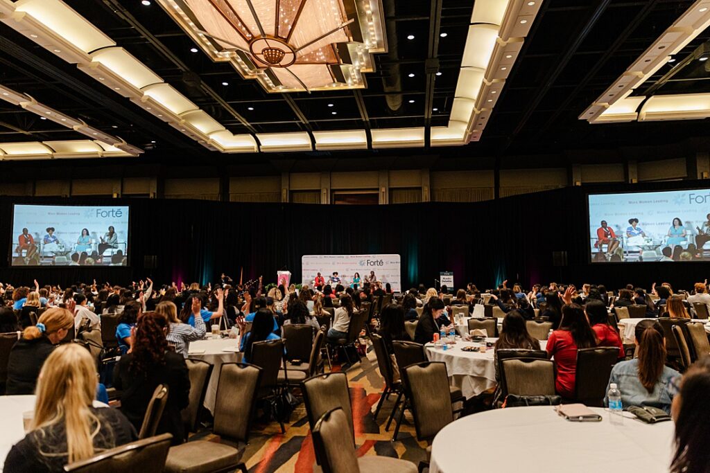 A wide shot of a ballroom during the Forte Foundation's MBA Women's Leadership Conference in Dallas, Texas. The foreground shows many circular ballroom tables with hundreds of women in professional business attire. The background shows a large stage with four women holding a discussion for the audience. Their image is being projected up on two large screens on either side of the stage.
