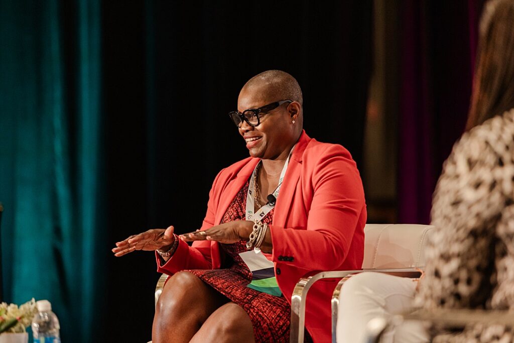A woman smiling and talking on stage during a panel at the Forte Foundation's MBA Women's Leadership Conference in Dallas, Texas. This woman is wearing a red and black dress with a red jacket and glasses.