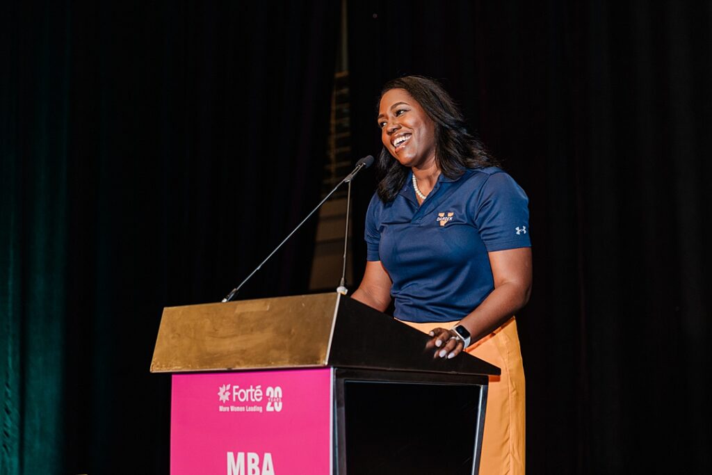 A woman talking on stage from a podium during the Forte Foundation's MBA Women's Leadership Conference in Dallas, Texas. She's wearing a branded polo representing her university.